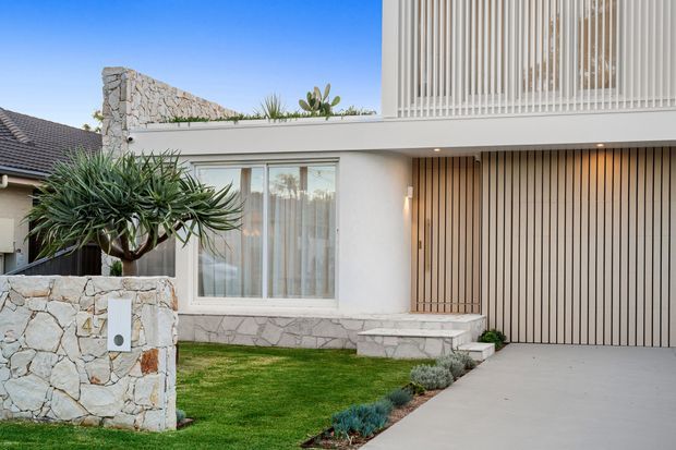 Contemporary two-storey residential facade with white rendered walls, a vertically slatted upper-level screen and matching timber-toned garage door. The frontage features irregular pale stone walling and a low stone-clad planter, a manicured lawn, sculptural drought-tolerant planting, large glazed windows, and a concrete driveway under a clear blue sky.