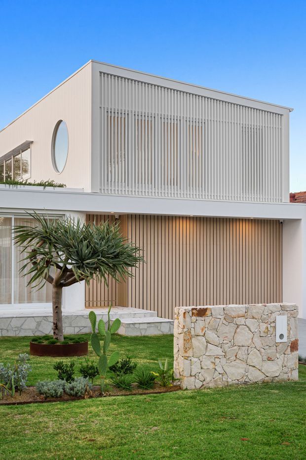 A contemporary two-storey residential facade in a light neutral palette. The upper level features a large screened facade of closely spaced vertical white fins, a distinctive oval window, and pale cladding. The lower level has warm vertical timber-look garage or entry screening, broad white horizontal slab edges, and expansive glazing. A landscaped front garden includes sculptural drought-tolerant planting, lawn, and a low natural-stone wall.
