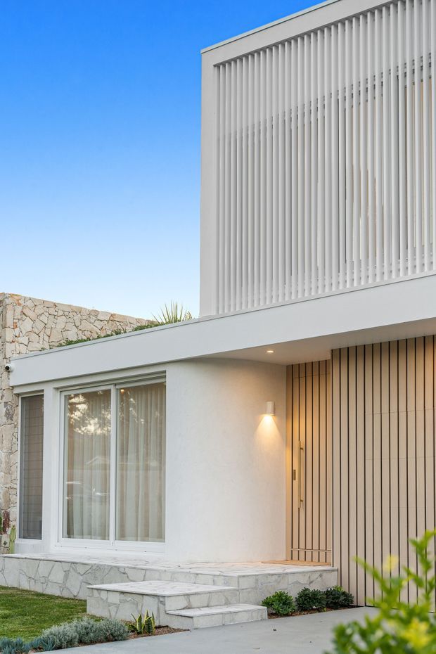 A contemporary residential facade viewed from outside, featuring a white rendered lower level, a projecting upper volume screened with closely spaced vertical white fins, a flat roofline, warm timber-toned vertical entry cladding, pale stone retaining and stair surfaces, and large glazed windows. Low planting and a paved entry path sit in front of the home under a clear blue sky.