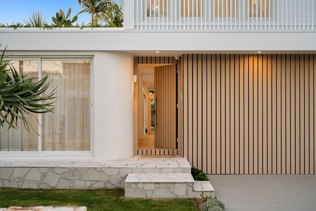A contemporary residential facade viewed from the front, featuring a white rendered upper structure, vertical white balcony screening, a warm timber-slatted garage or entry facade, and a pale irregular stone-clad plinth with steps. Tropical planting and a small lawn frame the entrance.