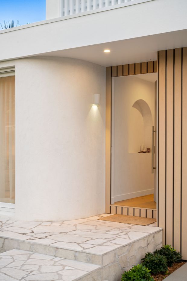 Contemporary residential entrance with a recessed glass front door framed by vertical timber battens. The entry is approached by broad pale natural-stone steps and paving, with white rendered walls, integrated exterior lighting, and a partially visible upper balcony or screen. Warm timber flooring and a softly lit arched interior niche are visible through the doorway.