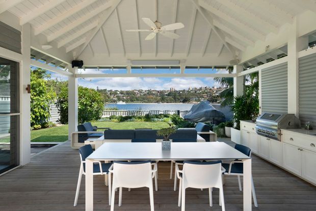 A bright covered outdoor entertaining area with a high white pitched timber ceiling, exposed rafters, ceiling fan, dining table and chairs, lounge seating, and a built-in outdoor kitchen with barbecue. The open sides overlook landscaped grounds and a waterfront beyond.