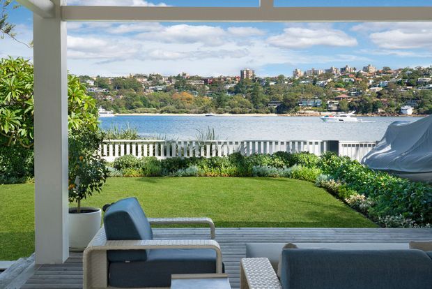 View from a covered outdoor terrace across a manicured lawn and white balustrade toward a broad waterfront, with residential hills and trees beyond. Outdoor lounge furniture sits in the foreground beneath a white-framed roof structure, creating a relaxed, open-air entertaining space.