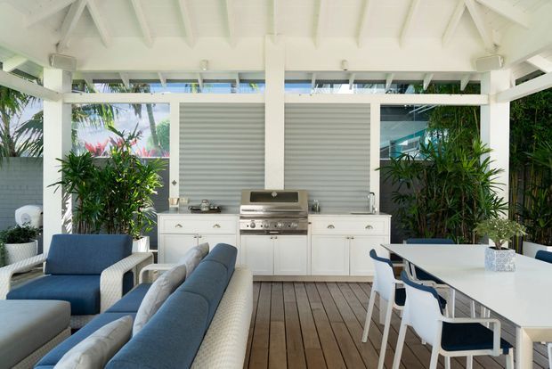 A bright, open-air covered entertaining area with a white-painted timber pitched roof and exposed rafters. The space combines a built-in outdoor kitchen with stainless-steel barbecue, white cabinetry and grey ribbed backsplash, alongside a dining table and blue-and-white lounge seating. Timber decking, tropical planting and large openings create a relaxed resort-like atmosphere.