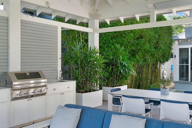 Bright covered outdoor entertaining area with a built-in stainless-steel barbecue, white cabinetry, dining table and chairs, and blue-and-white lounge seating. The space is framed by white structural beams and surrounded by dense tropical screening plants, with a pool or water feature visible beyond the dining area.