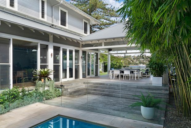 A bright residential courtyard viewed from beside a swimming pool, featuring a two-storey light-grey weatherboard house with white trim, extensive glazed doors and windows, a covered timber deck, outdoor dining area, steps, and tropical planting. The deck and pool create a relaxed indoor-outdoor entertaining setting.