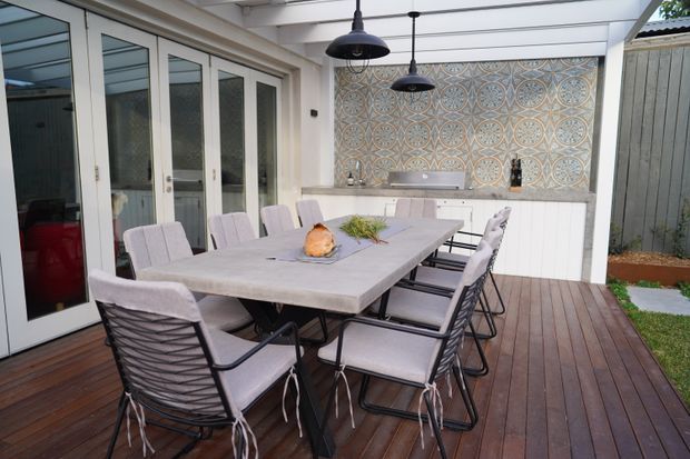Covered outdoor dining area on a timber deck, featuring a long dining table with upholstered chairs, a white built-in outdoor kitchen or bar cabinet, patterned tiled splashback, black pendant lights, and glazed folding doors opening to the house.
