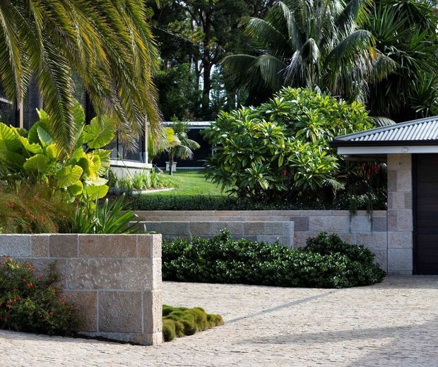Exterior view of a landscaped residential property with lush tropical planting, layered low stone retaining walls, and a pale textured paved driveway or forecourt. A single-storey structure with a grey metal roof and dark garage door is visible on the right.
