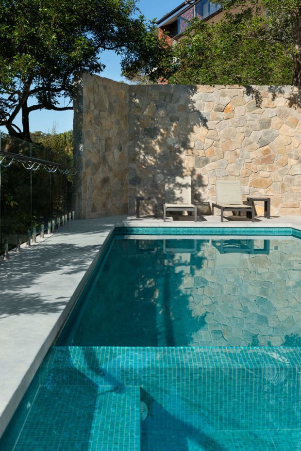 Sunlit outdoor swimming pool beside a pale stone-clad privacy wall, with turquoise mosaic pool tiling, concrete paving, glass balustrade, lounge chairs, mature trees and part of a modern multi-storey house visible above the wall.