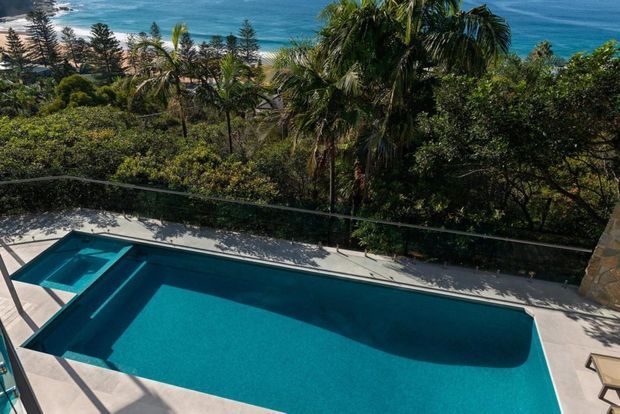 Elevated outdoor view of a large rectangular swimming pool surrounded by pale paving, with dense tropical landscaping and glimpses of a blue ocean coastline beyond. A stone-clad wall or column is visible at the right edge.
