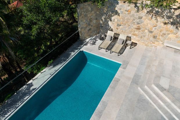 Elevated view of a rectangular swimming pool beside a broad pale stone-paved terrace. Two sun loungers sit against a rustic irregular-stone feature wall, with steps descending into the pool area and dense greenery surrounding the space.