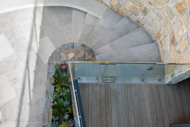 Top-down view of a curved spiral stairway with broad grey stone or concrete treads wrapping around a planted central feature. A rustic stacked-stone wall borders the stair, and a glass balustrade separates it from a dark timber-clad area.
