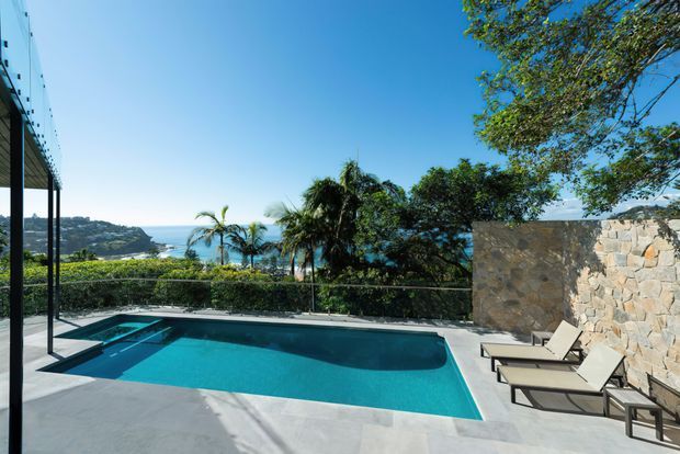 A modern outdoor pool terrace overlooking a bright coastal bay. The rectangular swimming pool is bordered by smooth grey paving, a glass balustrade, dark slender structural posts, and a natural irregular-stone feature wall. Lounge chairs sit beside the pool, with palms and coastal cliffs visible beyond.