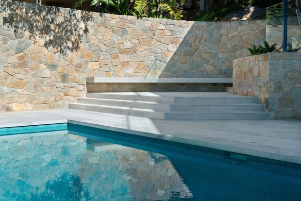 Outdoor swimming pool area with pale stone paving, broad steps, a built-in bench, and surrounding rough-cut natural stone retaining walls. Strong sunlight and reflected stonework create a warm, resort-like setting.