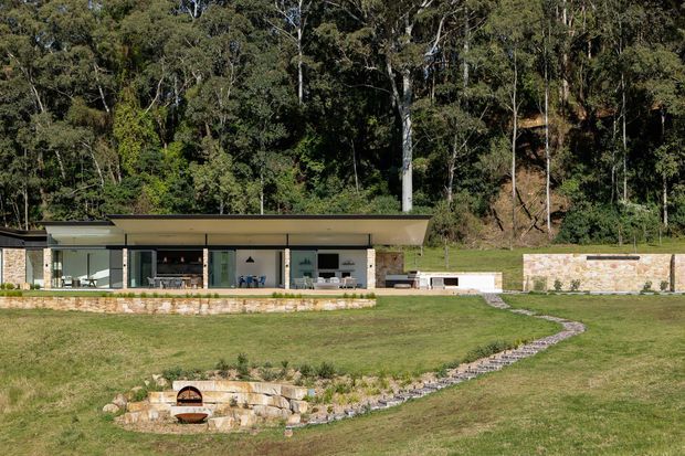 A low, single-storey modern pavilion set into a grassy, wooded site. The building has a broad flat roof with deep overhangs, extensive floor-to-ceiling glazing, pale stone-clad walls, and an outdoor entertaining area with a built-in fireplace and paved path. Dense native bush forms the backdrop.