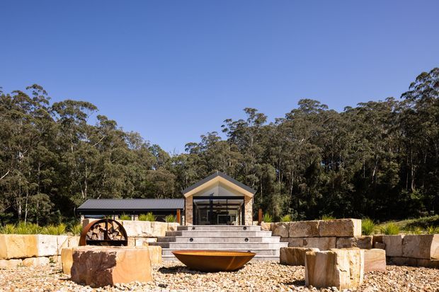 A small modern pavilion-like building set within a landscaped bush setting. The structure has a simple gabled roof, dark metal cladding on the side wing, warm timber or wood-toned detailing around the central glazed entrance, broad concrete steps, and substantial rough-cut stone retaining walls and boulders. A large decorative water wheel sits to the left, with pale gravel and sandstone-like blocks in the foreground.