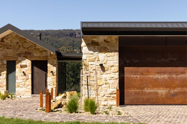 Exterior view of a low-profile property with rough natural-stone walls, a dark pitched roof over the smaller entrance wing, and a large rust-toned metal garage door beneath a broad flat-roofed section. A textured stone-paved driveway and native-style planting sit in front, with wooded hills visible beyond.