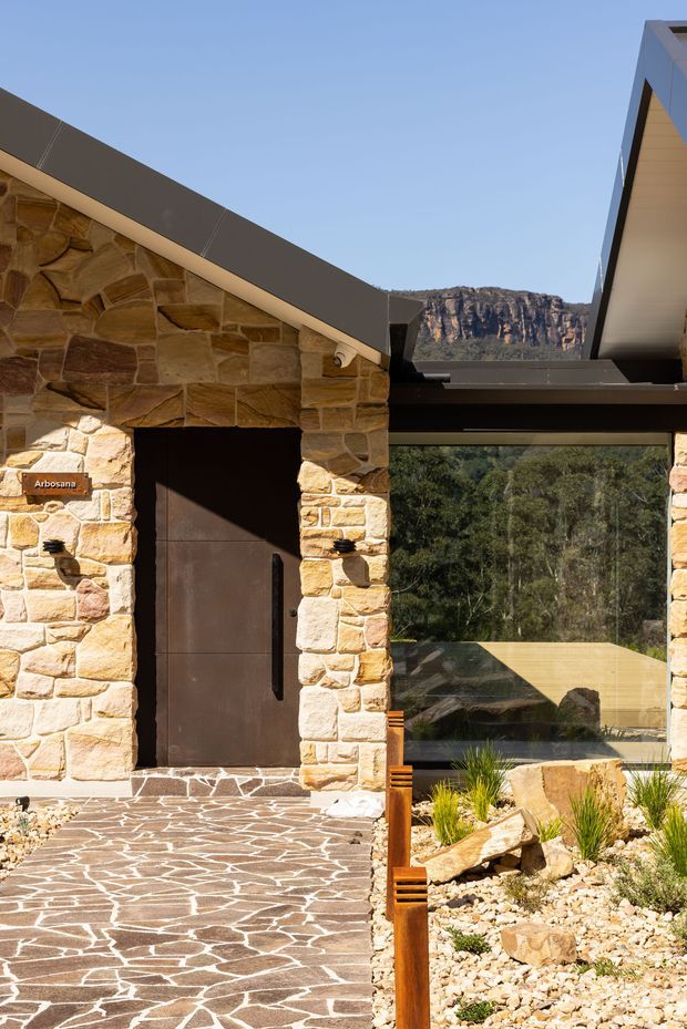 Exterior architectural detail of a contemporary low-profile building set against a dramatic rocky escarpment. The facade combines irregular warm sandstone masonry, dark metal roofing and framing, a large dark entry door, expansive glazing, and a patterned stone-paved approach. A dry landscaped garden with rocks and drought-tolerant planting is visible beside the entry.