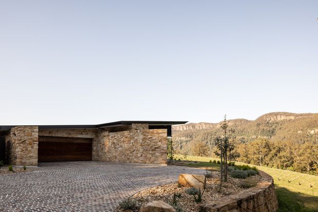 Low-profile contemporary house exterior with a broad flat roof, rough natural-stone walls, and an integrated double garage. A large cobbled driveway and stone-edged landscaped garden sit in front, with a wooded hilly landscape beyond.