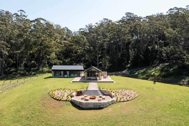 Aerial view of a spacious rural property set within dense eucalyptus woodland. A low, modern building with dark cladding and a pitched metal roof sits behind a landscaped lawn, with a central paved path leading to a circular stone outdoor fire pit and terraced garden beds.