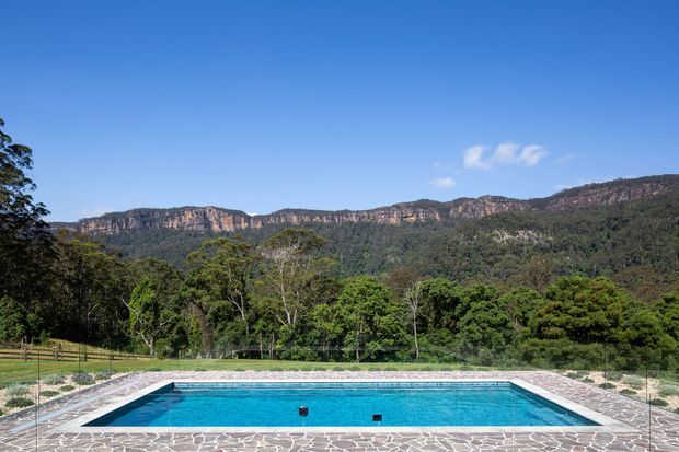 A rectangular outdoor swimming pool with bright blue water, surrounded by irregular stone paving and fencing. The pool sits in a landscaped rural setting with dense forest and dramatic sandstone escarpment mountains in the background under a clear blue sky.