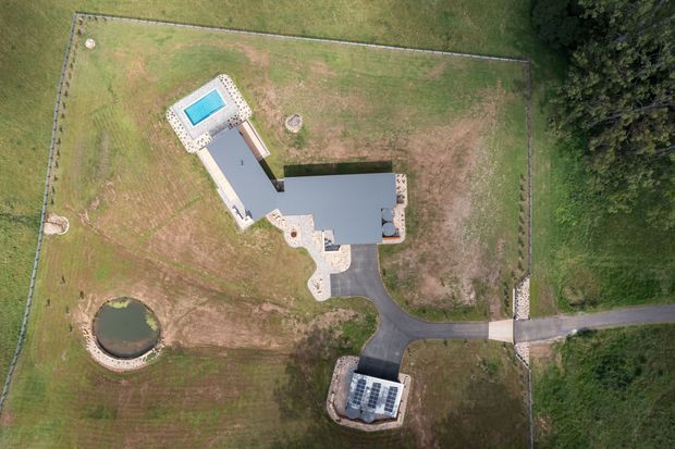 Aerial overhead view of a large rural property showing a contemporary building with a dark flat roof, pale paved terraces, a swimming pool, curved driveway, fenced grounds, landscaped lawn, and a small pond. Dense trees border the site.