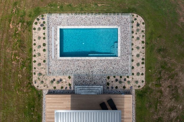 Aerial top-down view of a rectangular swimming pool surrounded by a patterned stone-paved terrace with curved corners and evenly spaced low plantings. A timber deck and covered outdoor structure extend from the lower edge, connected by steps.