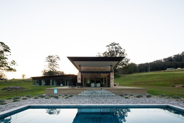 A contemporary single-storey pavilion-style house set in a landscaped rural setting, viewed across a swimming pool. The building features a dramatic projecting flat roof, extensive floor-to-ceiling glazing, dark horizontal cladding, stone-clad walls, broad concrete terraces and steps, and a symmetrical central entrance. Rolling lawns and trees surround the property.