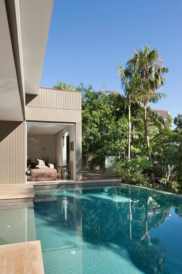 A contemporary residential exterior beside a clear swimming pool, with a partially open-plan living area visible through large glazed sliding doors. The building has smooth light-toned walls, vertical exterior cladding, broad roof overhangs, and a timber poolside deck. Lush tropical planting and palm trees surround the pool in bright sunlight.
