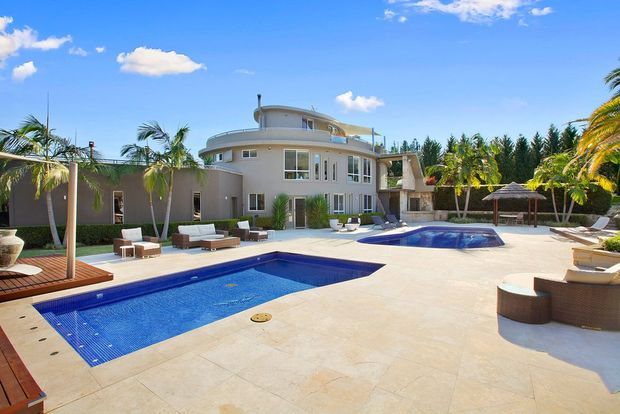 Bright exterior view of a large contemporary multi-level residence with curved roof and balcony forms, extensive pale paved terraces, two blue swimming pools, outdoor lounge furniture, palms, and landscaped screening vegetation. The scene has a resort-like tropical setting and strong indoor-outdoor entertaining focus.