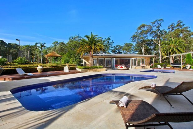 A sunny outdoor pool area with a large curved swimming pool surrounded by pale concrete paving, dark woven sun loungers, and a low modern single-storey pavilion. Tropical palms, mature trees, manicured hedges, a tennis court, and a small thatched structure form the landscaped setting.