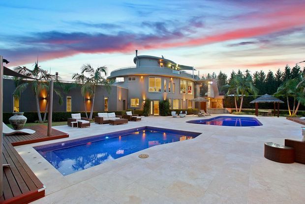 A large modern multi-level residence viewed from an expansive pool terrace at dusk. The property features rounded and rectilinear forms, extensive glazing, warm exterior lighting, multiple palm trees, a large swimming pool, a smaller raised spa or plunge pool, timber decking, pale paved surfaces, and outdoor seating.