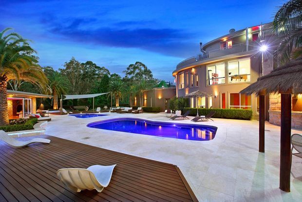 Twilight exterior view of a luxurious multi-storey residence centered around a large illuminated swimming pool. The property features curved contemporary architecture, extensive floor-to-ceiling glazing, warm interior lighting, a broad pale stone-paved pool terrace, dark timber decking, tropical planting, outdoor lounge furniture, and a thatched-roof pavilion.