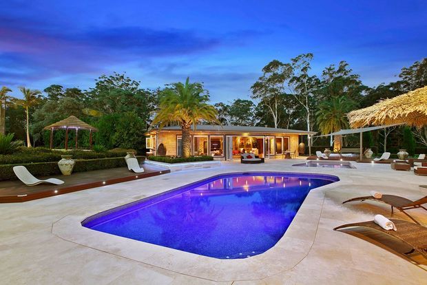 A landscaped resort-style outdoor pool area at dusk, with a large illuminated swimming pool, pale paved terrace, curved timber decking, sun loungers, tropical planting, thatched umbrellas, a gazebo, and a low modern pavilion with warm interior lighting.