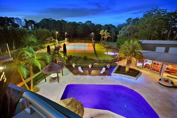 Elevated dusk view of a landscaped outdoor entertainment area with a curved illuminated swimming pool, poolside loungers, tropical planting, a thatched shade structure, and a fenced tennis court beyond. A low modern building with large windows and a metal roof borders the pool terrace.
