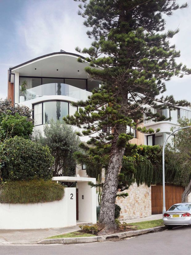 Street-facing view of a multi-storey contemporary residence with curved balconies, expansive glazing, white rendered walls, timber garage door, and a pale stone boundary wall. Mature landscaped planting and a large conifer partially screen the facade.