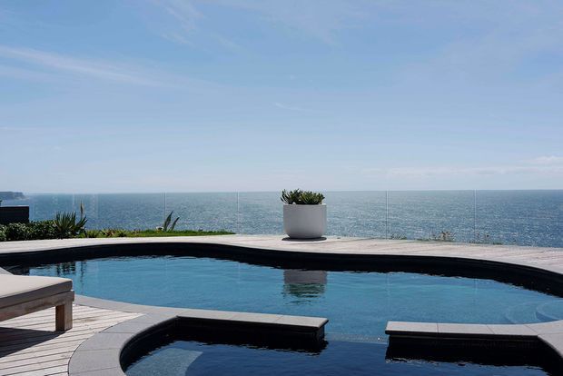 A modern infinity-edge swimming pool overlooking the ocean, with dark pool coping, pale paved decking, built-in bench-like pool ledges, and sparse potted and planted greenery. The scene is bright, open, and coastal.