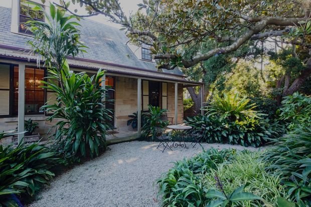 A leafy courtyard garden viewed from outdoors, featuring a traditional-looking house with a steep grey tiled roof, covered verandah, timber-framed openings, pale masonry walls, gravel paving, mature trees, dense tropical planting, and a small outdoor table and chairs.