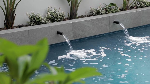 Close-up view of a modern swimming pool with two wall-mounted water spouts creating streams and ripples in the blue water. A raised grey tiled planter wall with tropical and leafy planting runs along the pool edge, while blurred foliage frames the foreground.