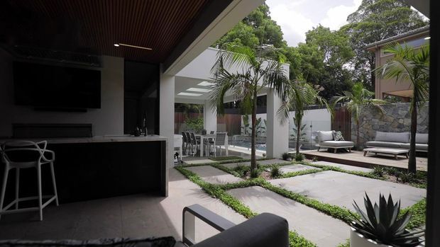 A modern indoor-outdoor entertaining area opening onto a landscaped courtyard. The shaded interior includes a dark kitchen or bar counter with stools, television and timber slatted ceiling, while the courtyard features large concrete stepping slabs, low planted borders, palms, sculptural planting, white columns and a pool or water feature. An outdoor lounge area sits against a stone-clad wall.
