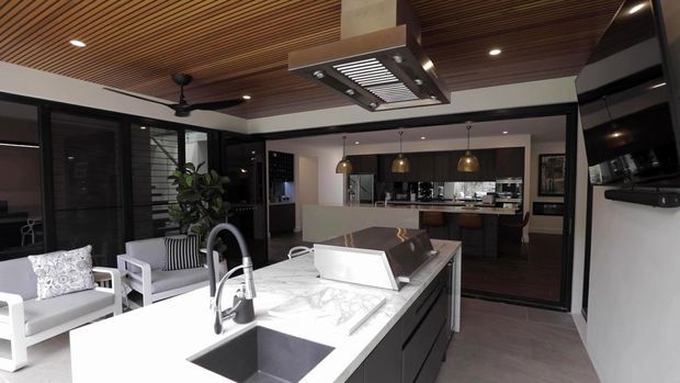 Open-plan contemporary kitchen viewed from a bright living or outdoor-adjacent area, featuring a large white marble-look island with integrated sink and dark cabinetry. Black-framed glazing and doors separate the kitchen from the adjoining space, while a timber slatted ceiling, suspended rangehood, pendant lights, and a wall-mounted television add strong architectural and functional details.