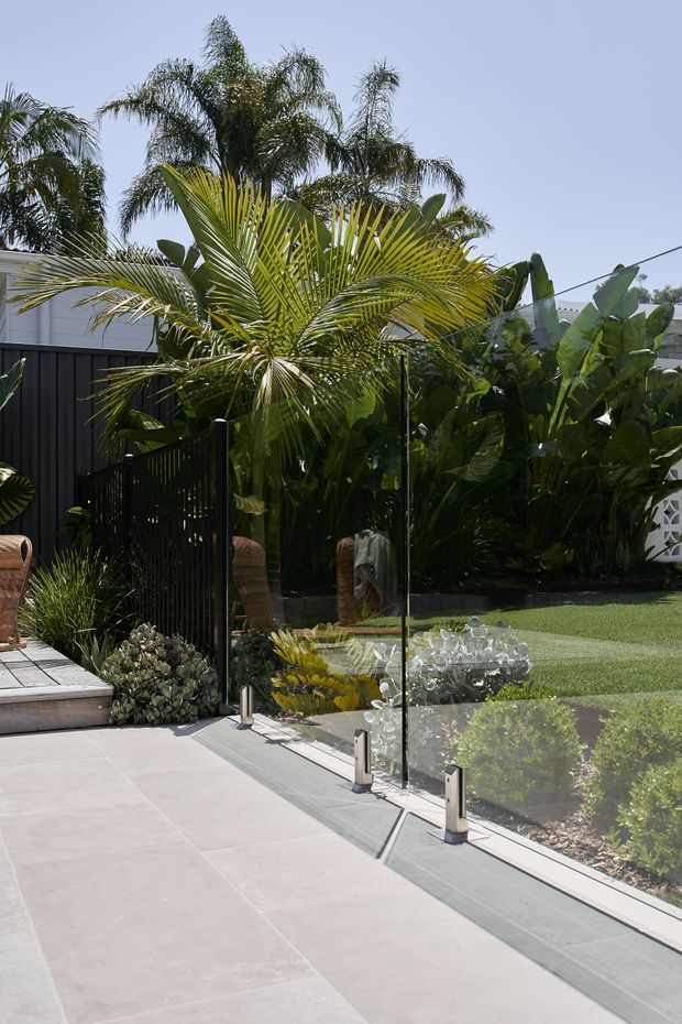 A sunny landscaped outdoor terrace or patio with large pale stone-format paving, frameless glass balustrade panels, and dark vertical metal fencing. Lush tropical planting, palms and broad-leaf foliage surround a manicured lawn, with partial outdoor seating visible at the left.