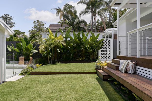 A bright landscaped residential courtyard or backyard with manicured lawns, lush tropical planting, and a raised timber deck. The white weatherboard-style house has an outdoor built-in bench with cushions, broad steps, white structural posts, and a decorative breeze-block privacy screen. Glass fencing and a small pool or water feature are visible at the left.