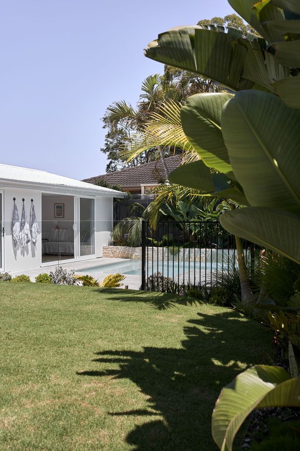 A bright residential backyard with a manicured lawn, tropical foliage, and a fenced swimming pool. A white pavilion or poolside room with large sliding glass doors opens toward the pool, with a glimpse of a bedroom or lounge inside. The scene has a relaxed tropical resort-like character under clear daylight.