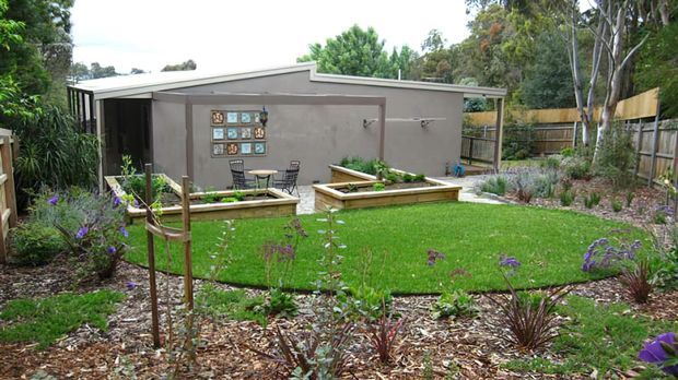 A landscaped backyard with a compact contemporary pavilion or garden room. The building has a low-pitched skillion roof, dark grey rendered walls, extensive glazing at one end, and an outdoor terrace with raised garden beds and seating. Native-style planting, lawn, timber fencing, and established trees surround the structure.