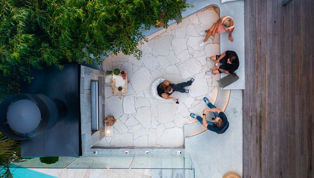 Overhead view of a landscaped outdoor courtyard or terrace with irregular pale stone paving, a built-in dark outdoor fireplace or structure, timber decking along one side, and several people gathered around outdoor seating. A glass-edged pool or water feature is partially visible at the bottom.