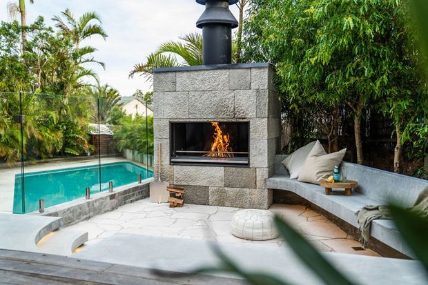 Outdoor entertaining courtyard with a built-in stone fireplace and chimney, curved concrete seating, pale stone paving, and a turquoise swimming pool enclosed by frameless glass balustrades. Lush tropical planting surrounds the space.