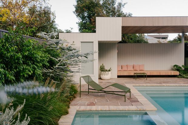 Contemporary landscaped pool courtyard with pale vertical-panel walls, a covered outdoor lounge featuring a built-in blush-toned bench, stone-paved terrace, and a second smaller pool or spa in the foreground. Dense planting softens the geometric architecture, while a sculptural green chaise lounge sits beside the water.