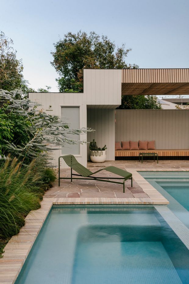 A refined contemporary courtyard pool area with two adjoining rectangular pools, pale stone paving, lush planting, and a modern outdoor entertaining pavilion. The pavilion features vertical light-toned cladding, a slatted roof and screen, built-in bench seating with cushions, and mid-century-inspired green outdoor loungers. The scene has a calm, warm, resort-like atmosphere.