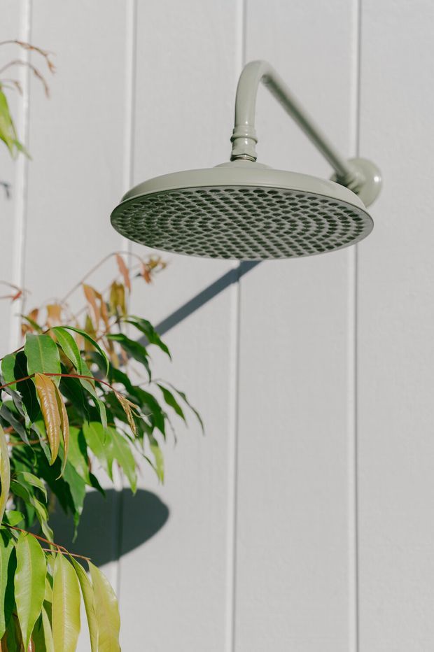 Close-up product-style image of a large round outdoor showerhead and curved wall-mounted arm in a muted pale green finish. The shower fixture is mounted against vertically panelled light grey exterior cladding, with leafy planting in the foreground and strong sunlight casting shadows.